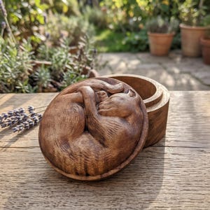 Puede incluir: Una caja redonda de madera con una tapa que presenta un relieve detallado de un gato durmiendo enroscado. La caja está hecha de madera de color marrón oscuro con un patrón de veta visible, mostrando al gato en posición de descanso.