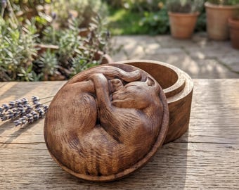 May include: A round wooden box with a lid featuring a detailed relief carving of a curled-up sleeping cat. The box is crafted from dark brown wood with a visible grain pattern, showing the cat in a resting position with its tail tucked in.