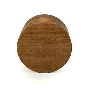 May include: A circular wooden box with a smooth, polished finish and a visible natural grain pattern. The lid is flat and fits flush with the base, creating a seamless cylindrical shape with warm, medium-brown tones throughout the entire object.