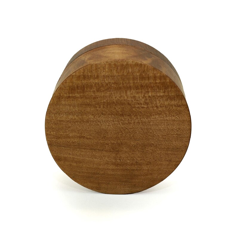 May include: A circular wooden box with a smooth, polished finish and a visible natural grain pattern. The lid is flat and fits flush with the base, creating a seamless cylindrical shape with warm, medium-brown tones throughout the entire object.