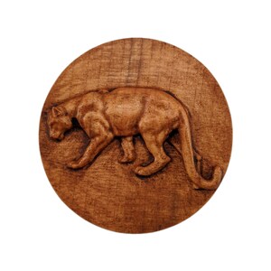 May include: A circular wooden plaque featuring a relief carving of a large cat in a prowling stance. The wood displays a natural medium brown grain with darker tones highlighting the muscular form of the feline as it moves across the surface.