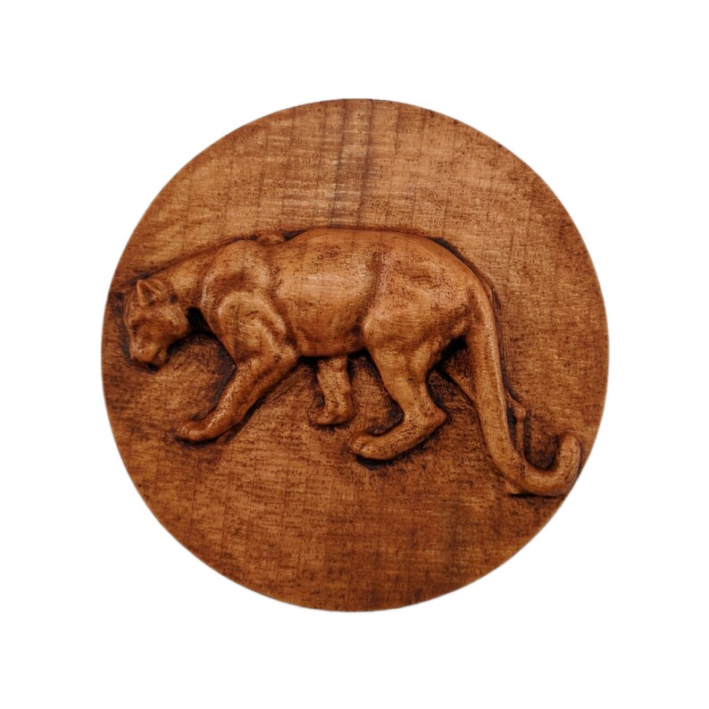 May include: A circular wooden plaque featuring a relief carving of a large cat in a prowling stance. The wood displays a natural medium brown grain with darker tones highlighting the muscular form of the feline as it moves across the surface.