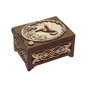 May include: A wooden music box with a hummingbird design. The box is dark brown with a light-colored hummingbird and floral design on the lid. The sides feature intricate, light-colored cut-out patterns. A small button is visible on the top.