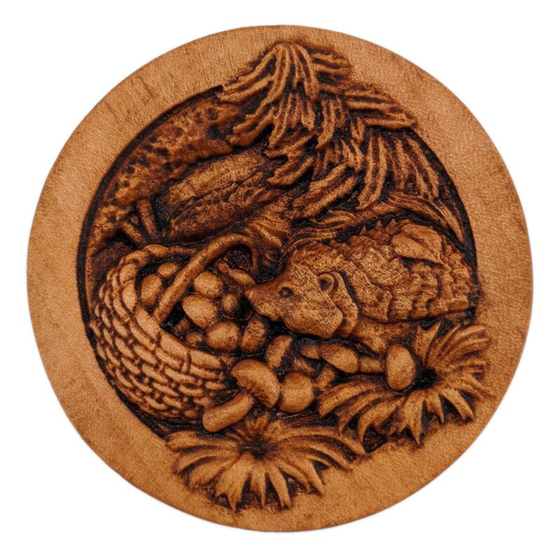 Round wooden box 3D carved with a hedgehog getting into a basket of mushrooms. Surrounded with flowers and leaves on the forest floor and eating the spilled mushrooms. Made from hard maple wood stained brown against a white background.