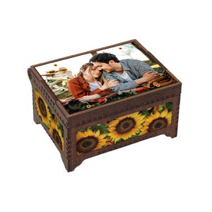 May include: A wooden keepsake box with a hinged lid. The box features a photograph on the lid and sunflower imagery on the sides. The box is brown with a rectangular shape and decorative edges.