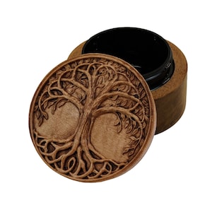 Puede incluir: Un recipiente redondo de madera con tapa extraíble que presenta una talla en relieve detallada de un árbol con ramas y raíces extendidas. La base del recipiente es de madera marrón y contiene un inserto circular negro y liso en el centro.