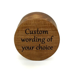 May include: A round, medium-brown wooden box with a smooth, polished finish. The top lid features dark, engraved text that reads, "Custom wording of your choice," centered in a clear, serif font against the natural wood grain pattern.