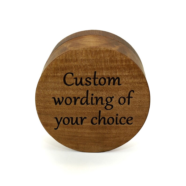 May include: A round, medium-brown wooden box with a smooth, polished finish. The top lid features dark, engraved text that reads, "Custom wording of your choice," centered in a clear, serif font against the natural wood grain pattern.