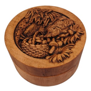 Round wooden box 3D carved with a hedgehog getting into a basket of mushrooms. Surrounded with flowers and leaves on the forest floor and eating the spilled mushrooms. Made from hard maple wood stained brown against a white background.