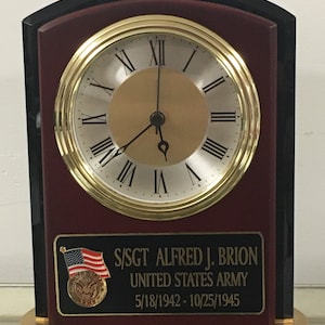 May include: A rectangular desk clock with a gold-tone clock face and Roman numerals. The clock has a gold-tone bezel and a dark red wooden frame. A black plaque displays text: S/SGT ALFRED J. BRION UNITED STATES ARMY 5/18/1942 - 10/25/1945.