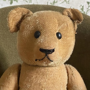May include: A vintage-style teddy bear crafted from tan, textured fabric. The bear features two round, dark-colored eyes and a hand-stitched black nose and mouth. It has two upright ears and a simple, rounded head shape with visible seams.