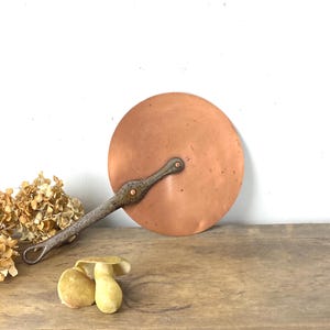 Puede incluir: Una tapa de sartén de cobre con un mango largo remachado. La tapa es redonda y tiene un aspecto ligeramente envejecido. El mango es de un metal más oscuro. La tapa de la sartén es probablemente para cocinar.