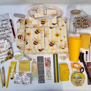 May include: A collection of stationery and lifestyle items including a bee and sunflower patterned tote bag, spiral notebooks with inspirational text, a yellow tumbler, a glass cup, bookmarks, pens, a small yellow wallet, and various floral-themed accessories.