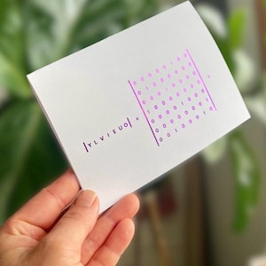 Peut inclure: Une carte blanche avec du texte violet et une grille de code binaire. La carte présente le texte "|YLVIEUO| x" sur le côté gauche. La grille de code binaire est sur le côté droit de la carte. La carte est tenue dans une main.