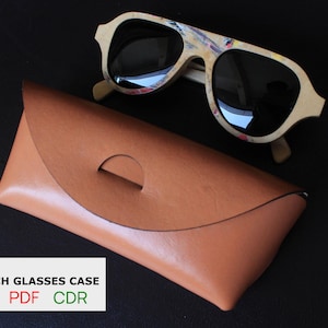 May include: A pair of sunglasses with a light-colored frame and dark lenses rests above a brown leather glasses case. The sunglasses frame has colorful accents. The glasses case has a curved design with a cutout. Text on the bottom reads "DIGITAL NO STICH GLASSES CASE SVG PDF CDR."