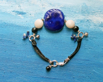 Pode incluir: Um bracelete de couro preto com uma conta de vidro azul e branca, detalhes metálicos prateados e contas de pedras preciosas pretas e azuis.