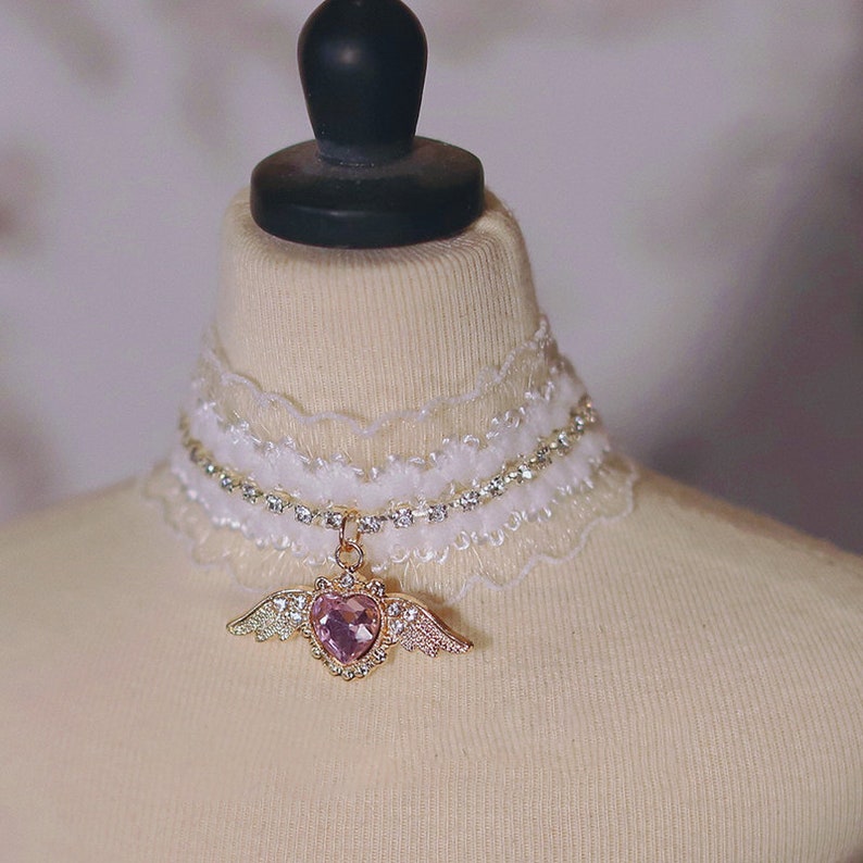 May include: A white lace choker with a rhinestone chain and a pink heart charm with gold wings.