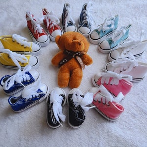 Könnte beinhalten: Eine Sammlung von 12 Miniatur-High-Top-Sneakern in verschiedenen Farben, darunter Gelb, Rot, Blau, Pink, Weiß und Schwarz. Die Sneaker sind in einem Kreis um einen braunen Teddybär mit einem braunen Band angeordnet.