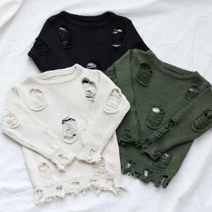 May include: Three distressed knit jumpers in black, white and green. The jumpers have a round neckline and long sleeves. The jumpers are made of a soft, comfortable material and have a distressed look with holes and frayed edges.