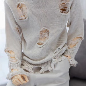 May include: A light beige sweater with distressed detailing and frayed edges. The sweater has several large holes throughout.