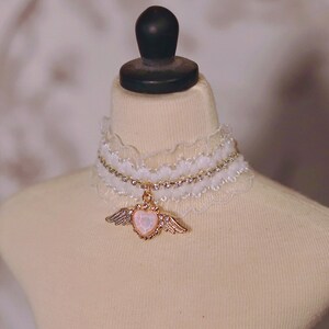 May include: A white lace choker with a rhinestone chain and a gold pendant featuring a heart with wings.