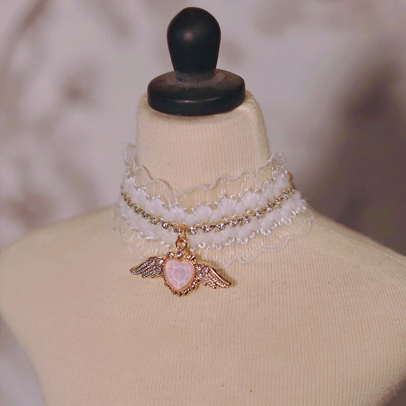 May include: A white lace choker with a rhinestone chain and a gold pendant featuring a heart with wings.