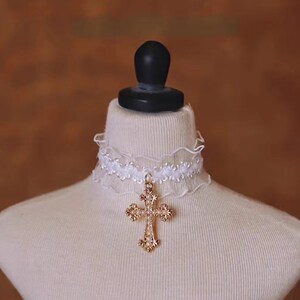 May include: White lace choker with a gold cross pendant. The cross is adorned with small white beads.