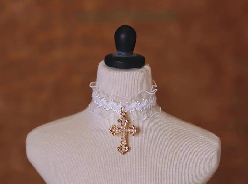 May include: White lace choker with a gold cross pendant. The cross is adorned with small white beads.
