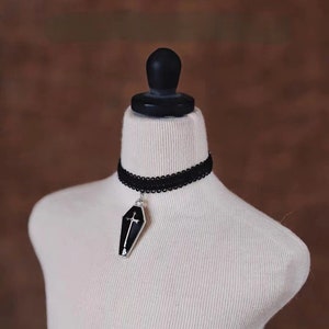 May include: A black choker necklace with a silver pendant shaped like a coffin with a cross on top.