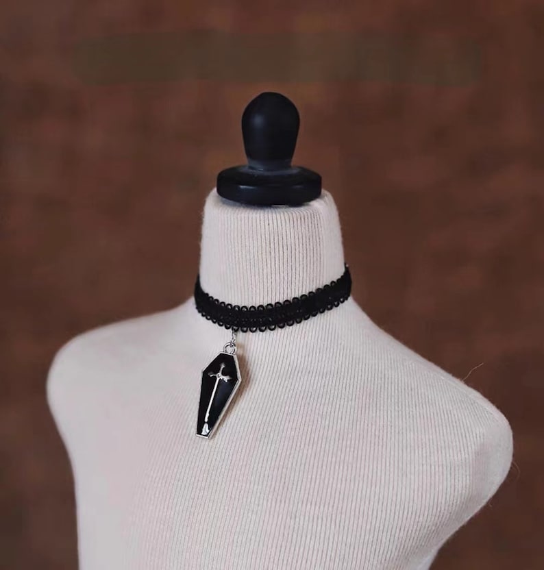 May include: A black choker necklace with a silver pendant shaped like a coffin with a cross on top.