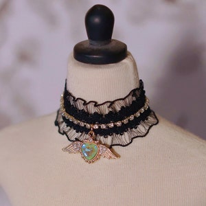 May include: A black lace choker with a rhinestone trim and a gold heart pendant with iridescent green wings.
