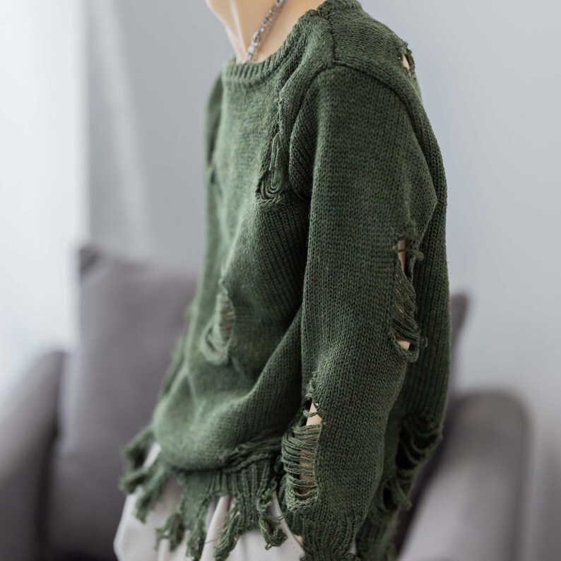 May include: A green distressed sweater with frayed edges and holes throughout. The sweater has a crew neck and long sleeves.