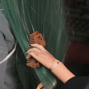 Puede incluir: Una mano sosteniendo un peine de madera con tallas intrincadas. El peine se est&aacute; utilizando para cepillar el cabello largo y verde.