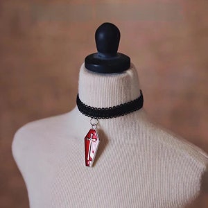 May include: A black choker necklace with a silver pendant shaped like a red coffin with a white cross on it.