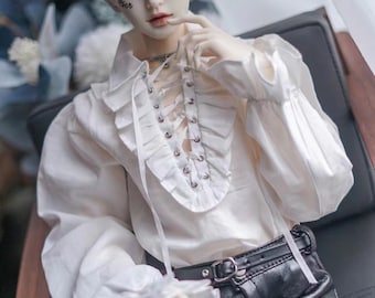 May include: A white doll with blonde hair wearing a white ruffled shirt with a lace-up front and black pants. The doll has a tattoo that says "oxer" on its face.