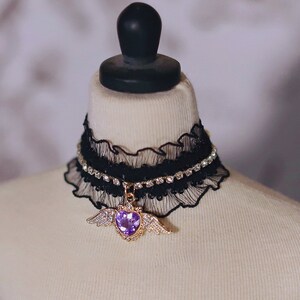 May include: A black lace choker with a rhinestone trim and a gold pendant featuring a purple heart with wings.