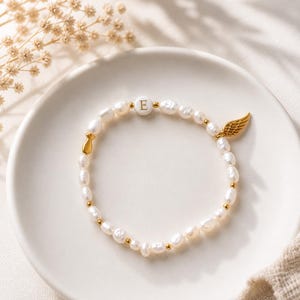 Può includere: Un braccialetto di perle con ciondoli dorati, tra cui un'ala e una perla con la lettera "E". Il braccialetto è presentato su un piatto bianco. Le perle sono bianche.