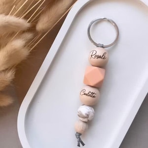 Könnte beinhalten: Ein Schlüsselanhänger aus Holzperlen mit den Namen "Rosalie" und "Calotta", die in die Perlen graviert sind. Der Schlüsselanhänger besteht aus natürlichen Holzperlen und einem silbernen Schlüsselring.