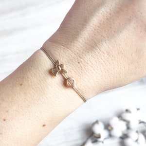 Puede incluir: Un brazalete de cordón fino marrón con un dije de ángel dorado y un dije de disco dorado con la letra "A" en él.