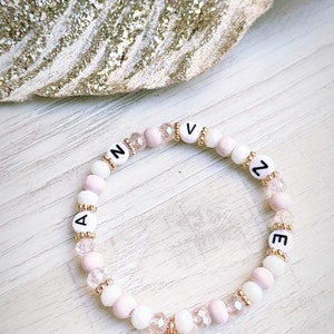 Könnte beinhalten: Ein rosa-weißes Perlenarmband mit einem goldenen Herz-Anhänger. Das Armband hat die Buchstaben "AVNZEE" darauf.