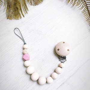 May include: A wooden pacifier clip with a light pink and white bead design. The clip features a round wooden clip, a series of wooden beads, a pink geometric bead, and a bead with the letter "R". The clip is attached to a gray cord.