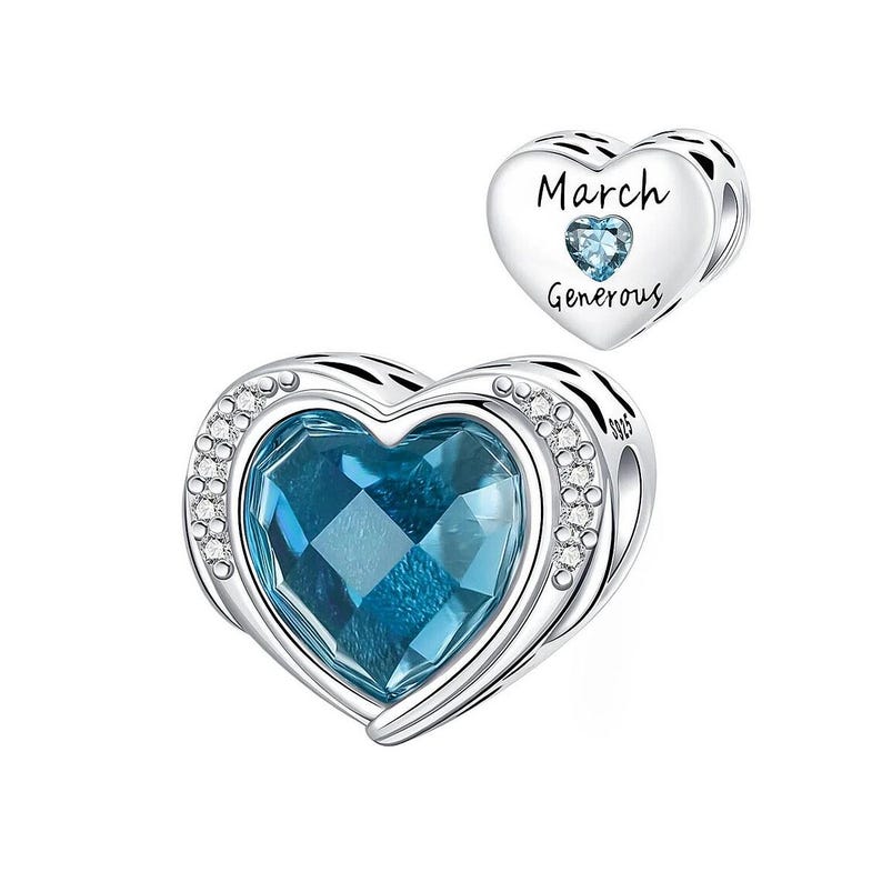 May include: A silver heart-shaped charm with a large, faceted blue gemstone center. The charm is accented with small clear crystals around the edge. A smaller heart charm with the words "March" and "Generous" is also visible.