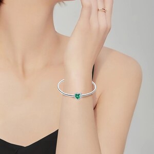 May include: A silver-toned bangle bracelet with a heart-shaped, emerald-green charm. The bracelet is a simple, open design, and the heart charm is centered on the band. The bracelet is worn on a wrist.