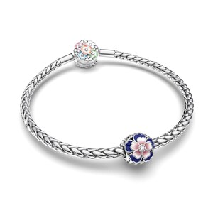 May include: A silver bracelet with a woven chain design. The bracelet features two flower charms. One charm has a rainbow of colors, and the other has a blue, pink, and white flower design. The bracelet is on a white background.