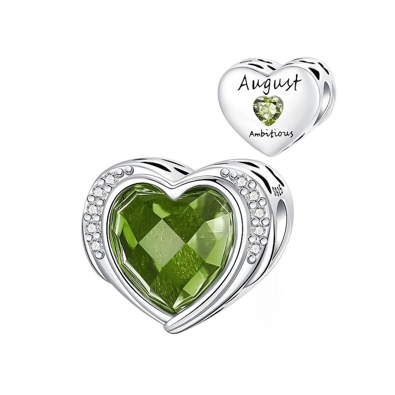 May include: A heart-shaped charm with a large, faceted green gemstone center, surrounded by a silver frame and small clear stones. The charm is labeled "August" and "Ambitious" with a smaller green stone.
