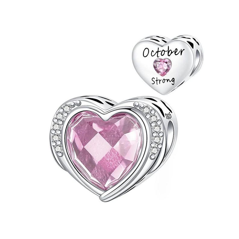 May include: A silver heart-shaped charm with a large pink gemstone center, surrounded by small clear crystals. A smaller heart charm with the words "October Strong" and a pink gemstone is also visible.