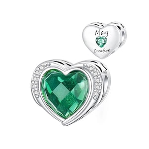 May include: A silver heart-shaped charm with a large, faceted green gemstone center, surrounded by small clear crystals. A smaller heart charm with the word "May" and "Creative" and a green gemstone is also shown.