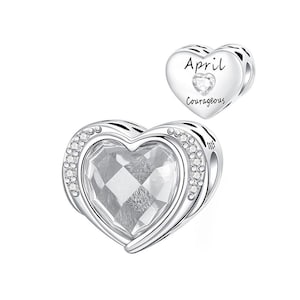 May include: A silver heart-shaped charm with a large, faceted clear crystal center, surrounded by a row of small clear crystals. A smaller heart charm with the word "April" and "Courageous" is also shown.