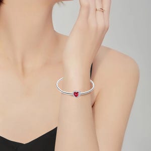 May include: A silver-colored bangle bracelet with a heart-shaped charm. The heart charm is red with a silver border. The bracelet is worn on a wrist.