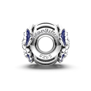 May include: A silver charm with blue enamel accents. The charm features the word "Manyilus" and the number "925". The design includes floral or leaf-like shapes. The charm is designed to be threaded onto a bracelet or necklace.